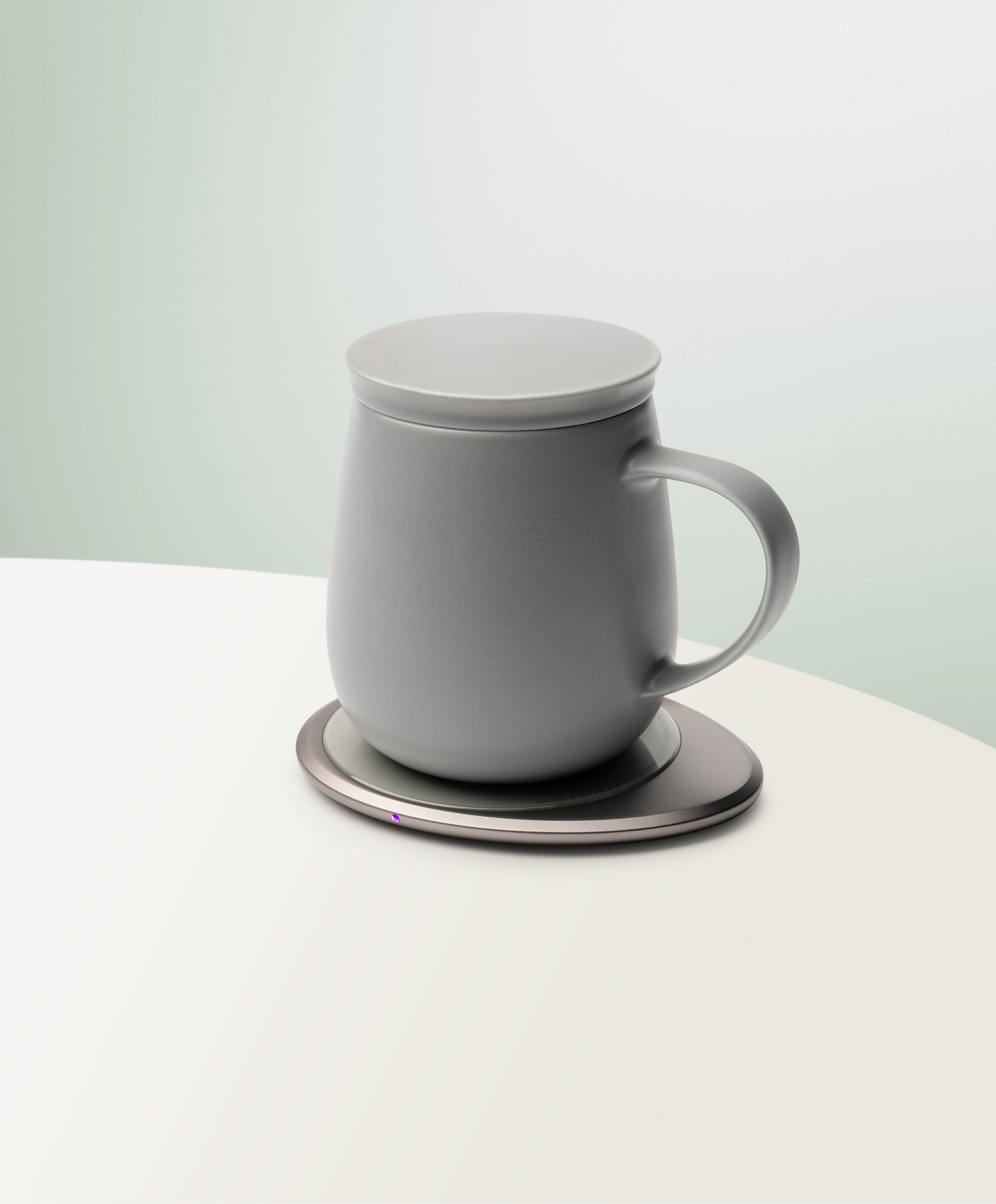 Ui 3 Self-Heating Ceramic Mug 12 fl oz / 355 mL in Soft Gray with lid angled top-side view on OHOM Charging Pad 3