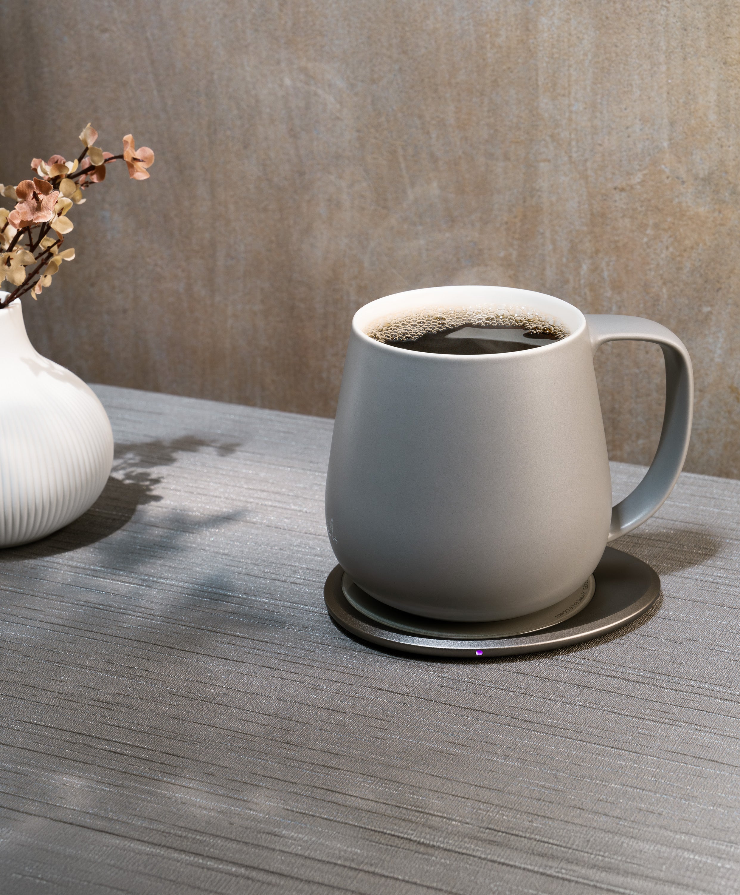 Ui+ self-heating ceramic mug set 18 fl oz / 532 mL in Soft Gray charging on OHOM Pad 3, filled with hot coffee and steam rising