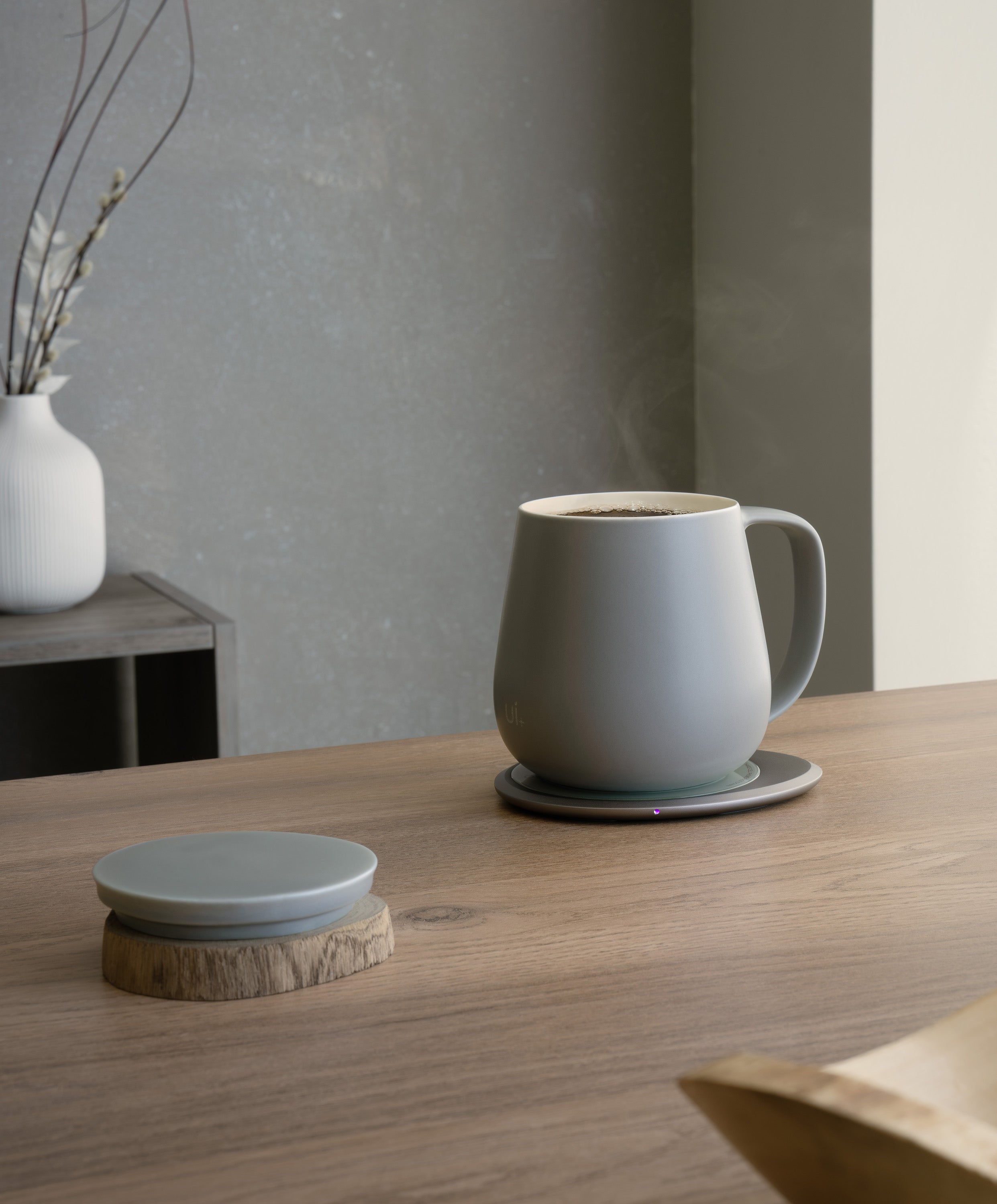 Ui+ self-heating ceramic mug set 18 fl oz / 532 mL in Soft Gray charging on OHOM Pad 3, lid placed on wooden coaster