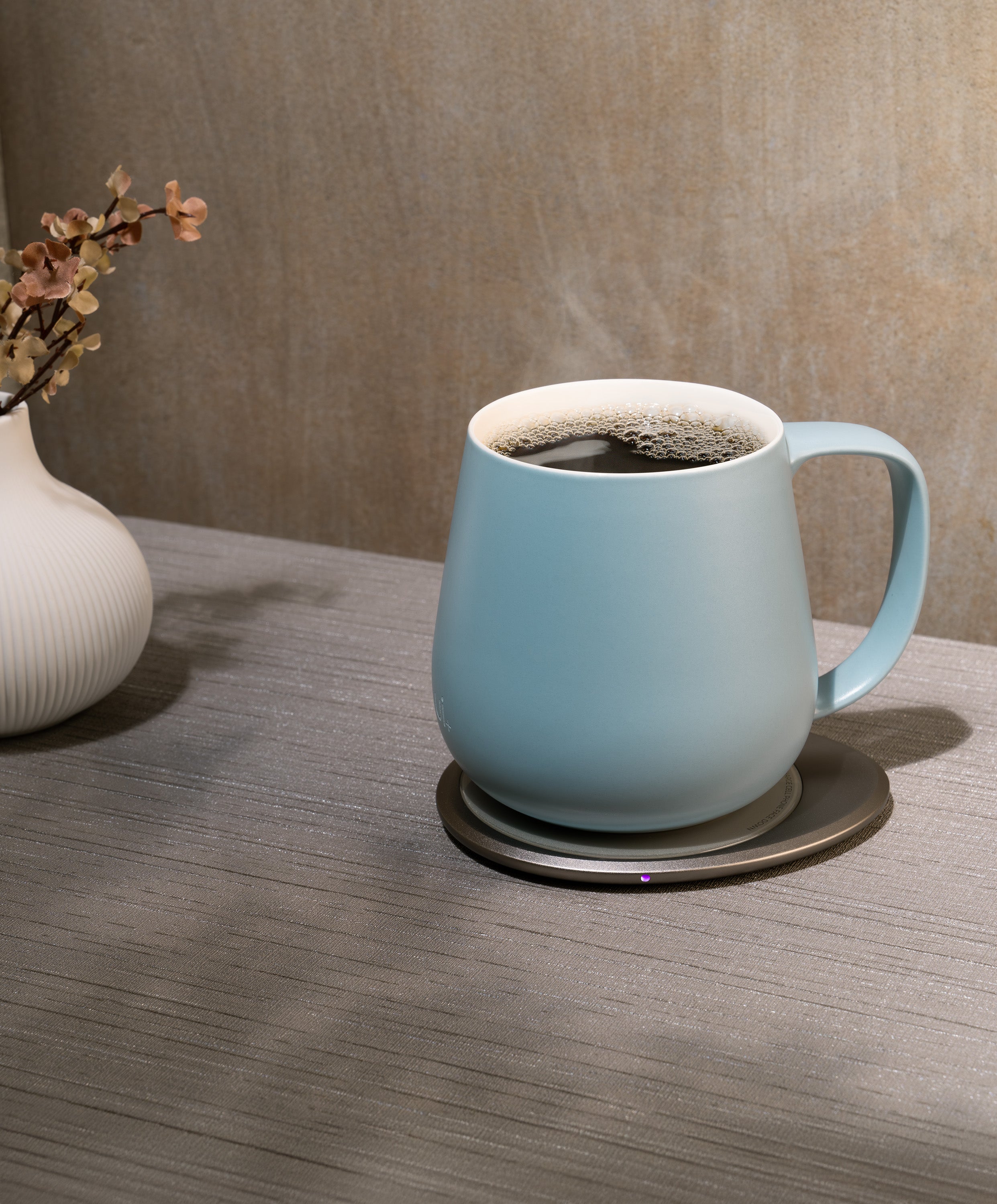 Ui+ self-heating ceramic mug set 18 fl oz / 532 mL in Moonmist Blue charging on OHOM Pad 3, filled with hot coffee and steam rising