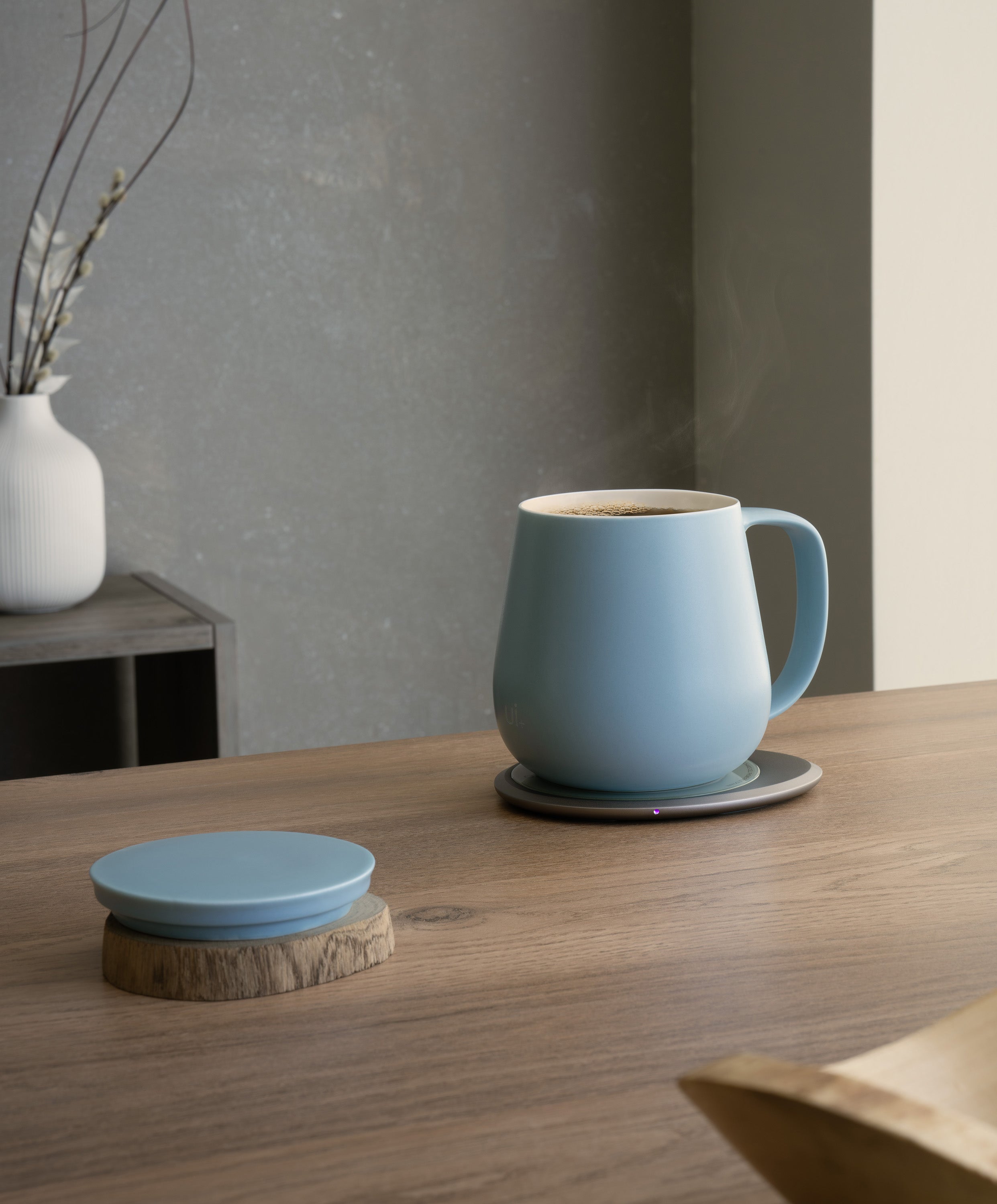 Ui+ self-heating ceramic mug set 18 fl oz / 532 mL in Moonmist Blue charging on OHOM Pad 3, lid placed on wooden coaster