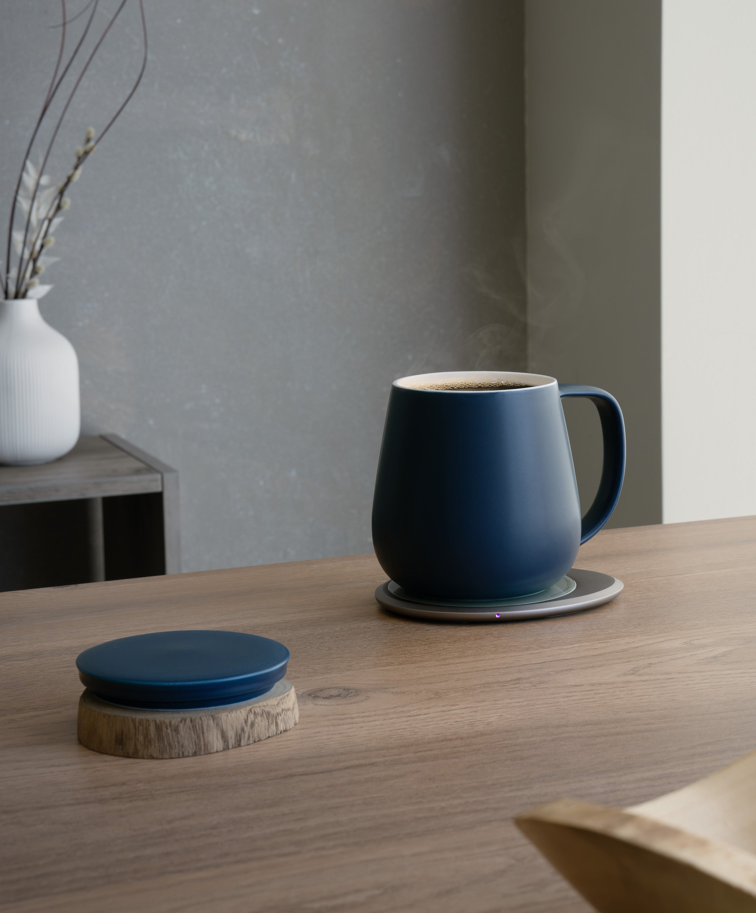 Ui+ self-heating ceramic mug set 18 fl oz / 532 mL in Deep Navy charging on OHOM Pad 3, lid placed on wooden coaster