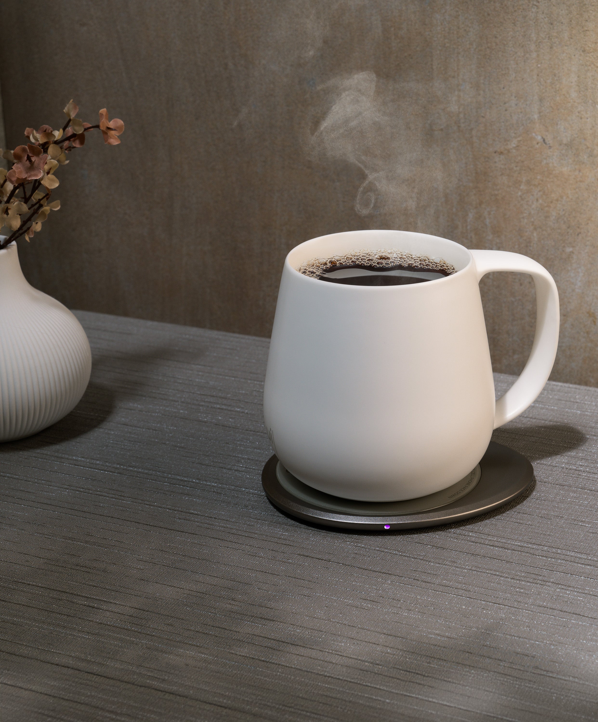 Ui+ self-heating ceramic mug set 18 fl oz / 532 mL in Jasmine White charging on OHOM Pad 3, filled with hot coffee and steam rising