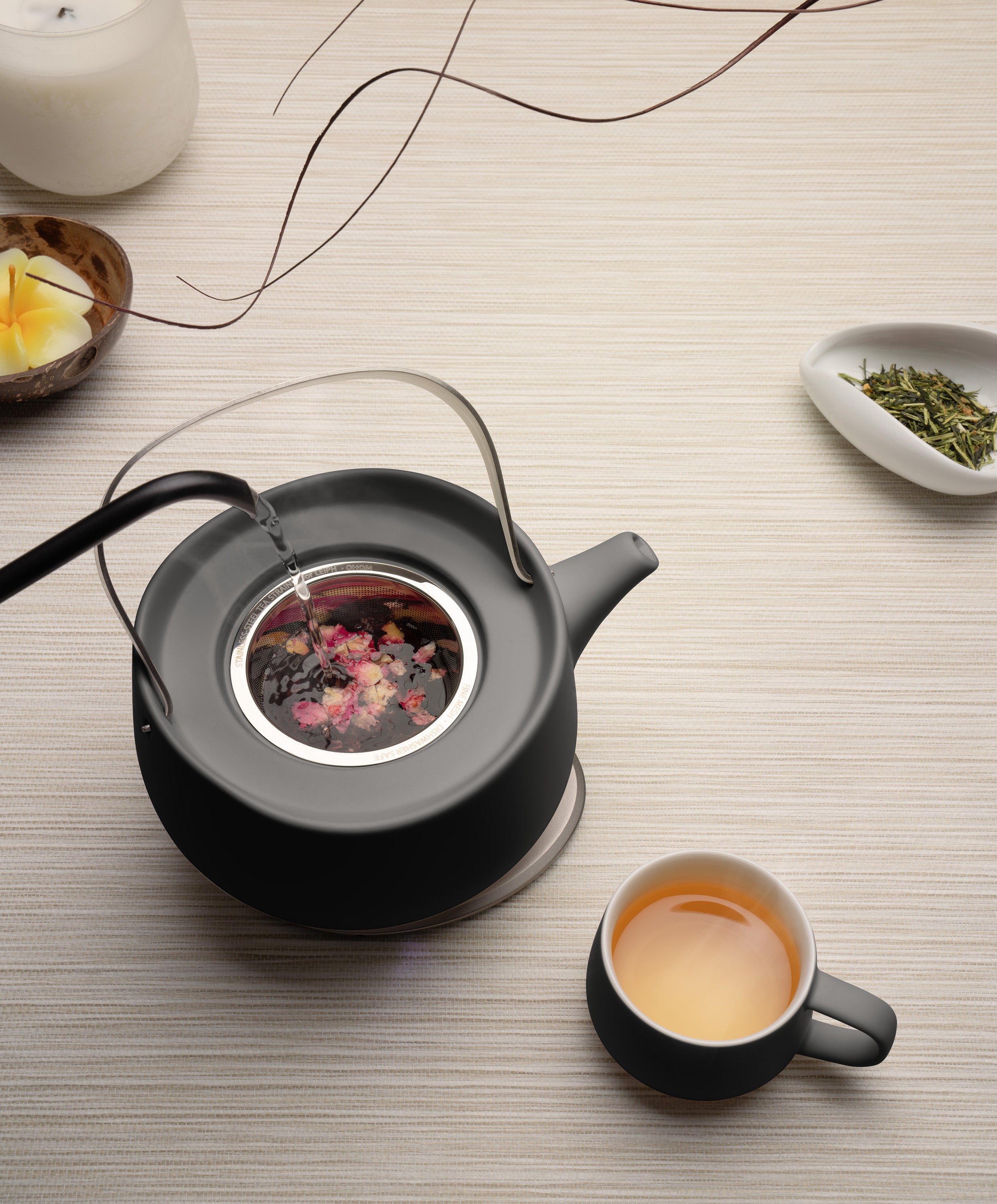 Leiph self-heating ceramic teapot 20 fl oz / 600 mL in Inkstone Black with herbs charging on OHOM Pad 3 as water is poured next to Ui mug with tea