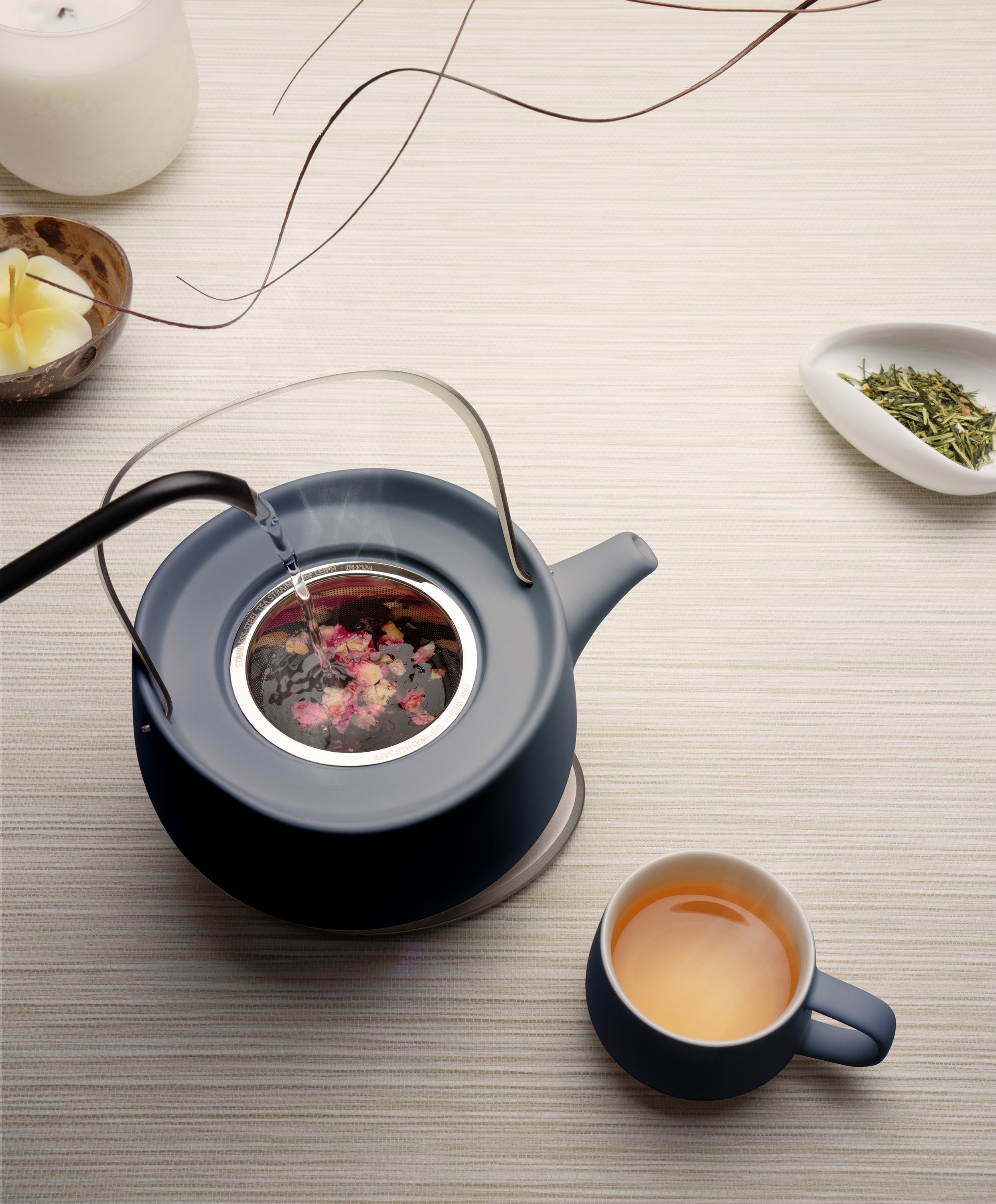Leiph self-heating ceramic teapot 20 fl oz / 600 mL in Deep Navy with herbs charging on OHOM Pad 3 as water is poured next to Ui mug with tea