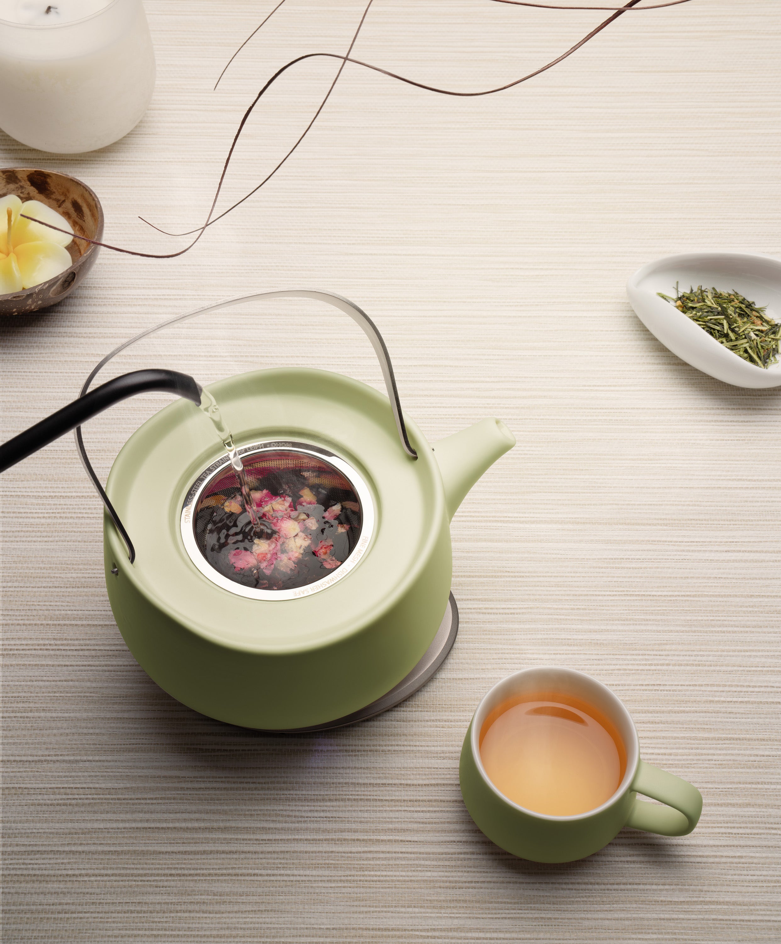 Leiph self-heating ceramic teapot 20 fl oz / 600 mL in Verde Green with herbs charging on OHOM Pad 3 as water is poured next to Ui mug with tea
