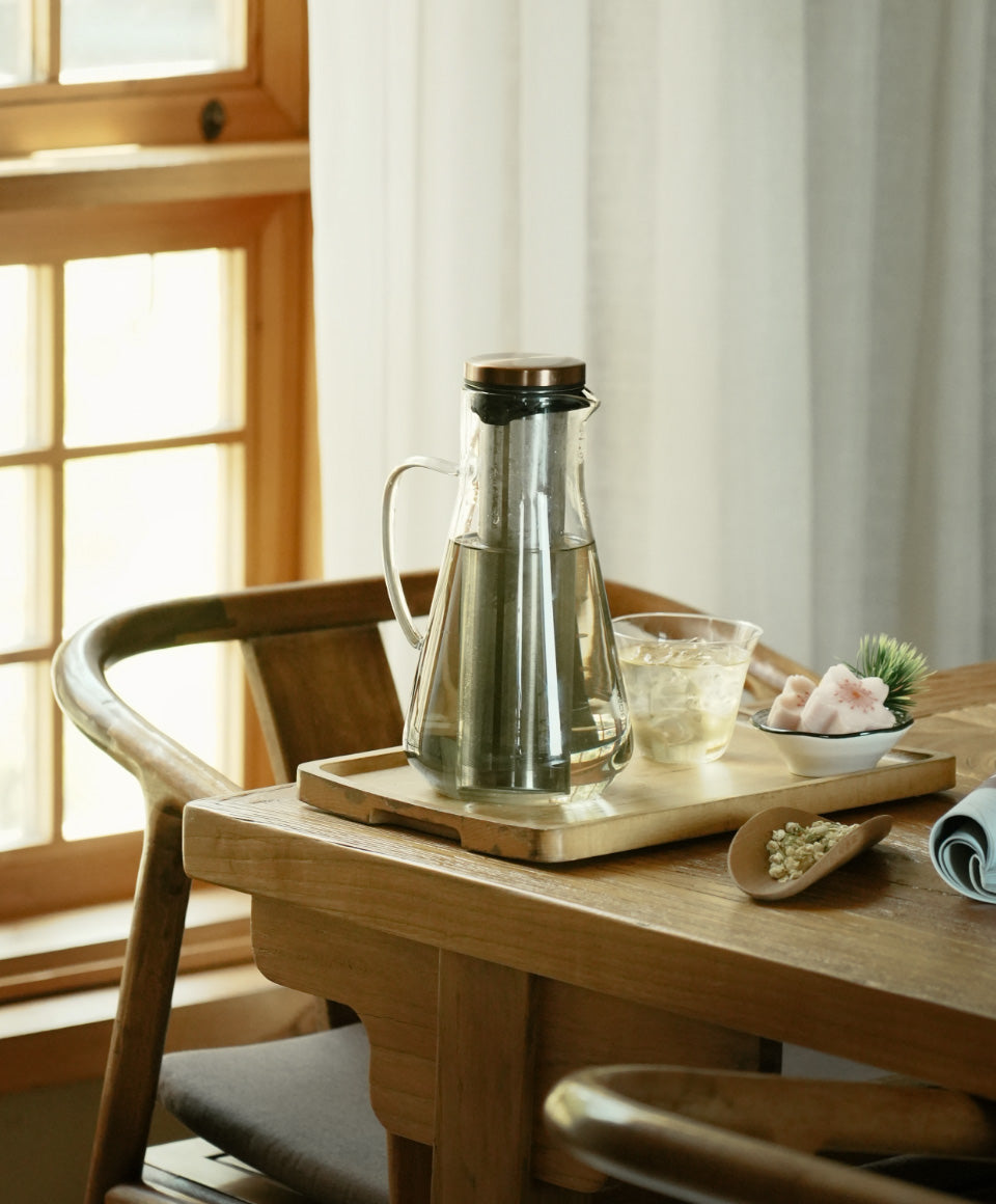 Sio cold-infusion pitcher 54 fl oz / 1600 mL handmade from borosilicate glass with copper lid on wooden tray, glass of brewed tea beside it