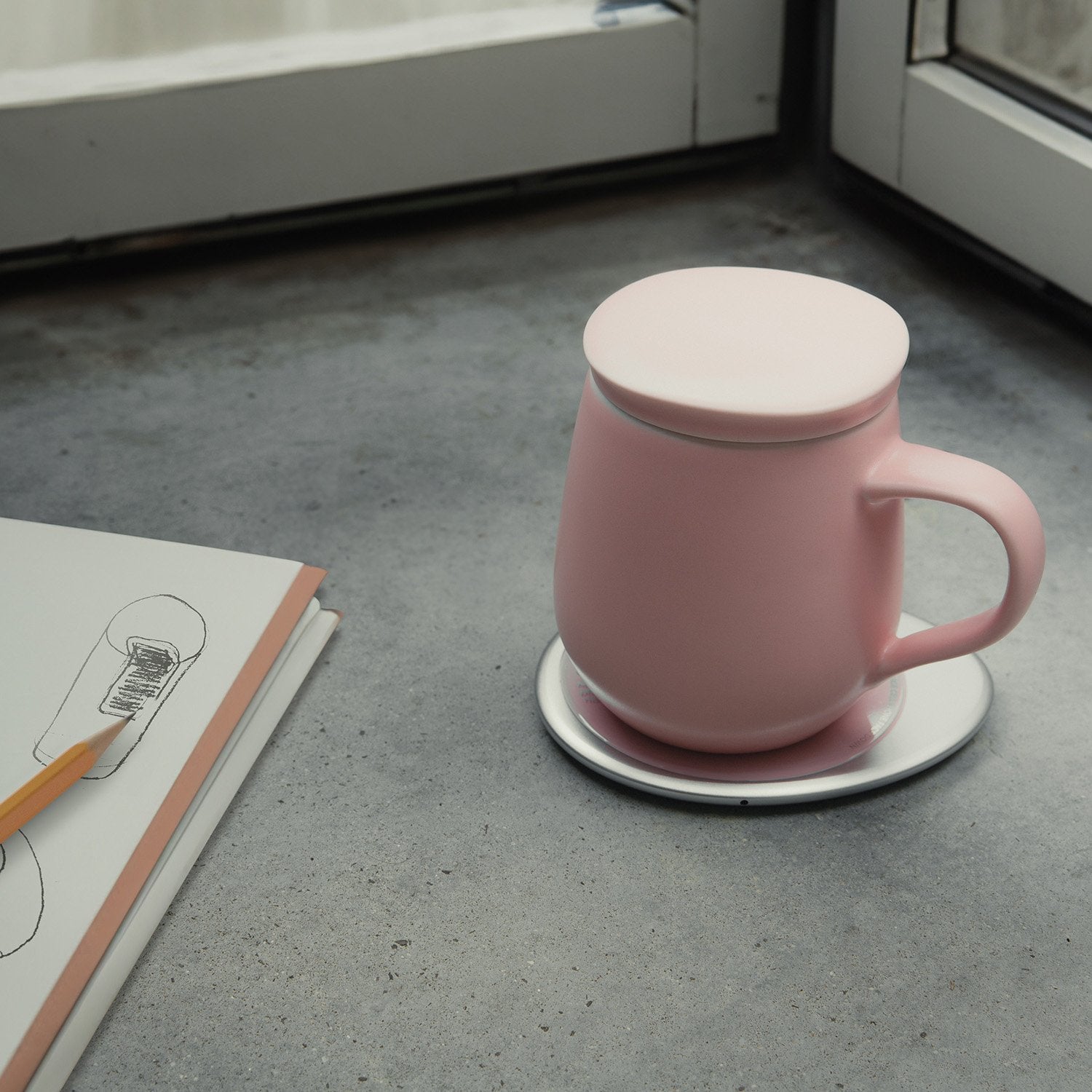 Pink mug with lid on heating pad next to drawing notebook